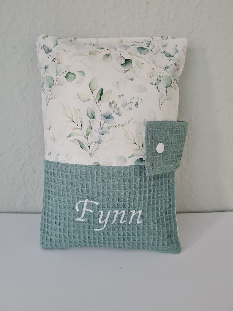 May include: A rectangular pillow with a floral pattern on the top half and a textured, light green bottom half. The floral pattern features green and blue leaves on a white background. The name "Fynn" is embroidered in white on the bottom half.