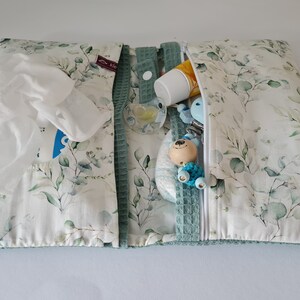 May include: A baby changing pad with a white and green eucalyptus leaf pattern. The pad is open, revealing a pack of wipes, a pacifier, a diaper, and other baby essentials. The pad has a green waffle trim and a zipper closure.