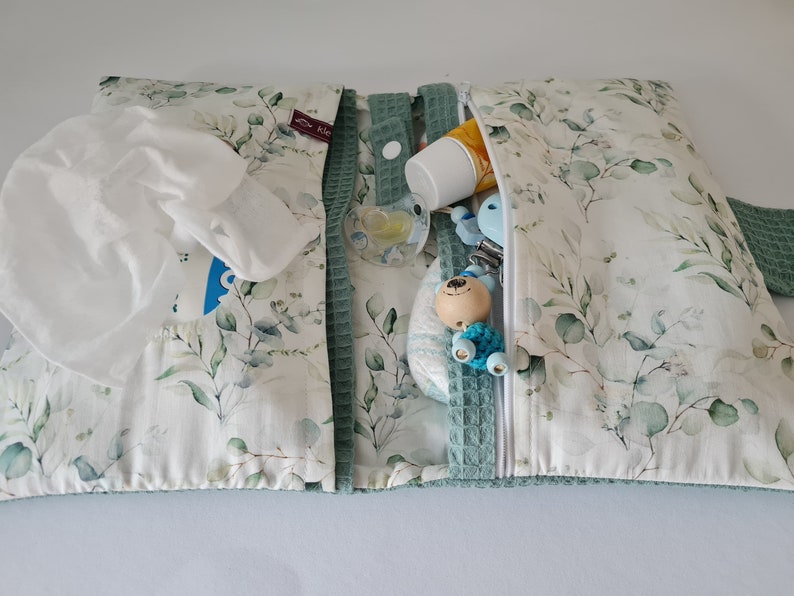 May include: A baby changing pad with a white and green eucalyptus leaf pattern. The pad is open, revealing a pack of wipes, a pacifier, a diaper, and other baby essentials. The pad has a green waffle trim and a zipper closure.