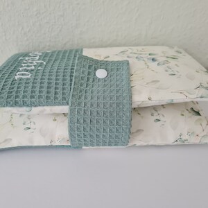 May include: Two decorative pillows. The top pillow is teal waffle weave with the name "Sophia" embroidered in white. The bottom pillow has a white background with a green and gray floral pattern. A teal waffle weave flap with a white button is attached.