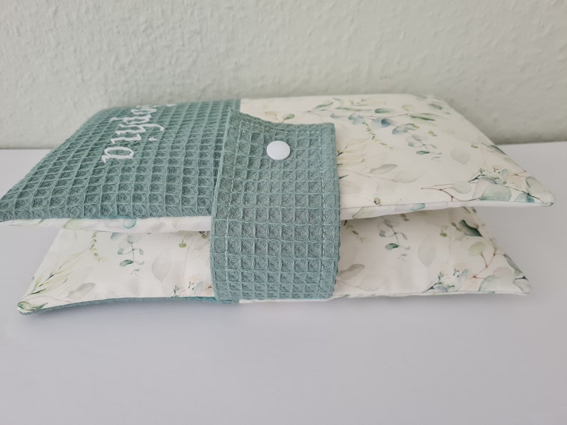 May include: Two decorative pillows. The top pillow is teal waffle weave with the name "Sophia" embroidered in white. The bottom pillow has a white background with a green and gray floral pattern. A teal waffle weave flap with a white button is attached.