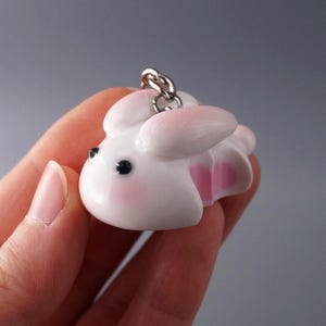 May include: A white bunny keychain with pink accents on the ears and inner body. The bunny has black eyes and rosy cheeks. A silver-colored chain is attached to the top of the bunny.