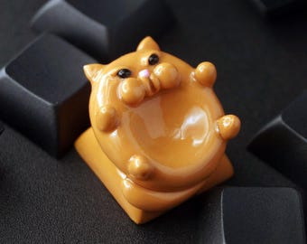 May include: A custom orange cat-shaped keycap with a glossy finish. The keycap features a rounded body, small paws, and a pink nose. It is surrounded by black keyboard keys, creating a contrast in color and texture.