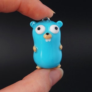 Puede incluir: Un pequeño dije de dibujos animados azul claro con un lazo plateado. Tiene ojos blancos grandes, una nariz marrón y pequeños brazos y pies marrones. El dije se sostiene entre dos dedos sobre un fondo oscuro.
