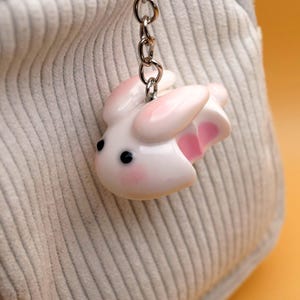 May include: A white bunny keychain with pink accents, including the ears and inner ears. The bunny has black eyes and rosy cheeks. The keychain is attached to a silver chain. The background is a light gray, corduroy-like fabric.