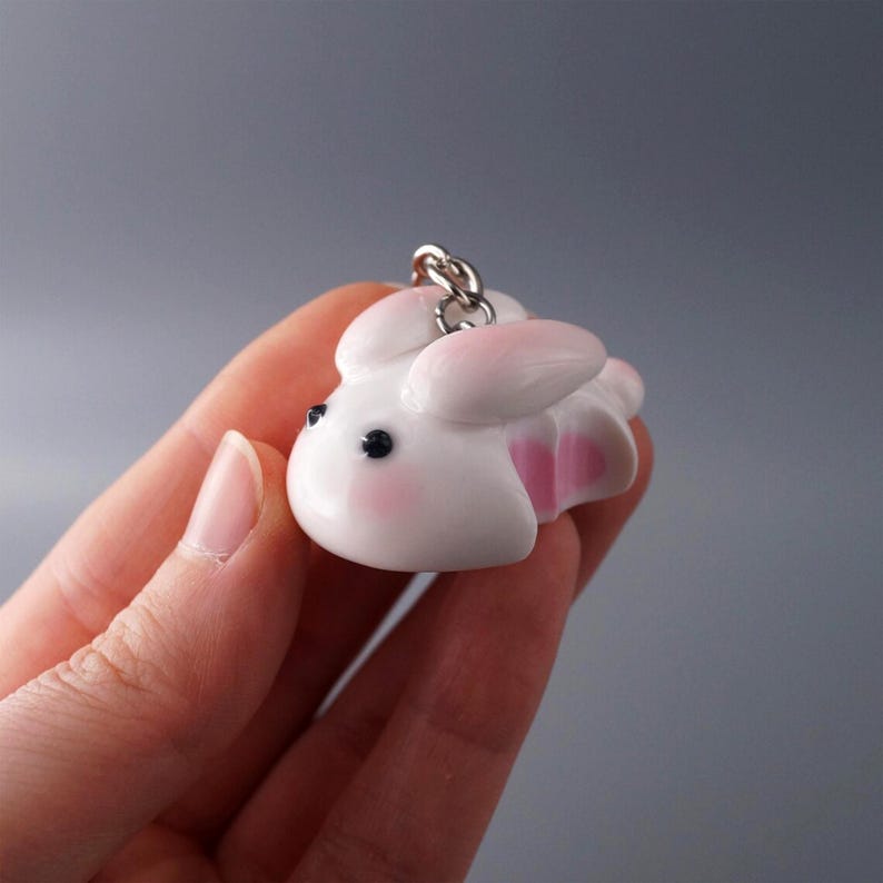 May include: A white bunny keychain with pink accents on the ears and inner body. The bunny has black eyes and rosy cheeks. A silver-colored chain is attached to the top of the bunny.