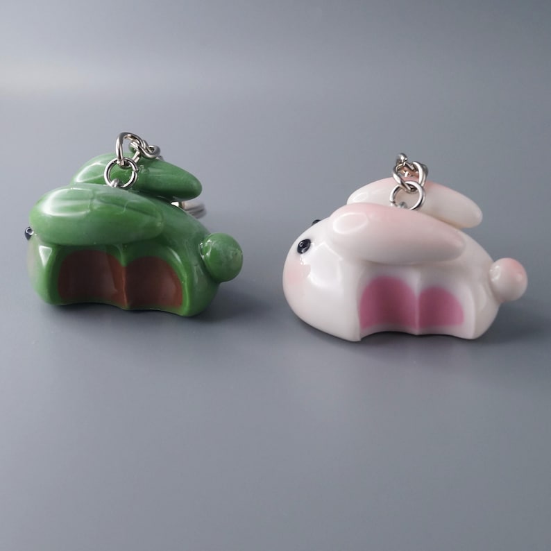 May include: Two ceramic bunny keychains. One is green with brown accents, the other is white with pink accents. Both have black eyes and silver key rings. The keychains are small, decorative, and likely for personal use.