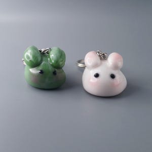 May include: Two keychains shaped like cartoon mice. One is green with black eyes, the other is white with pink accents. Both have silver key rings attached.