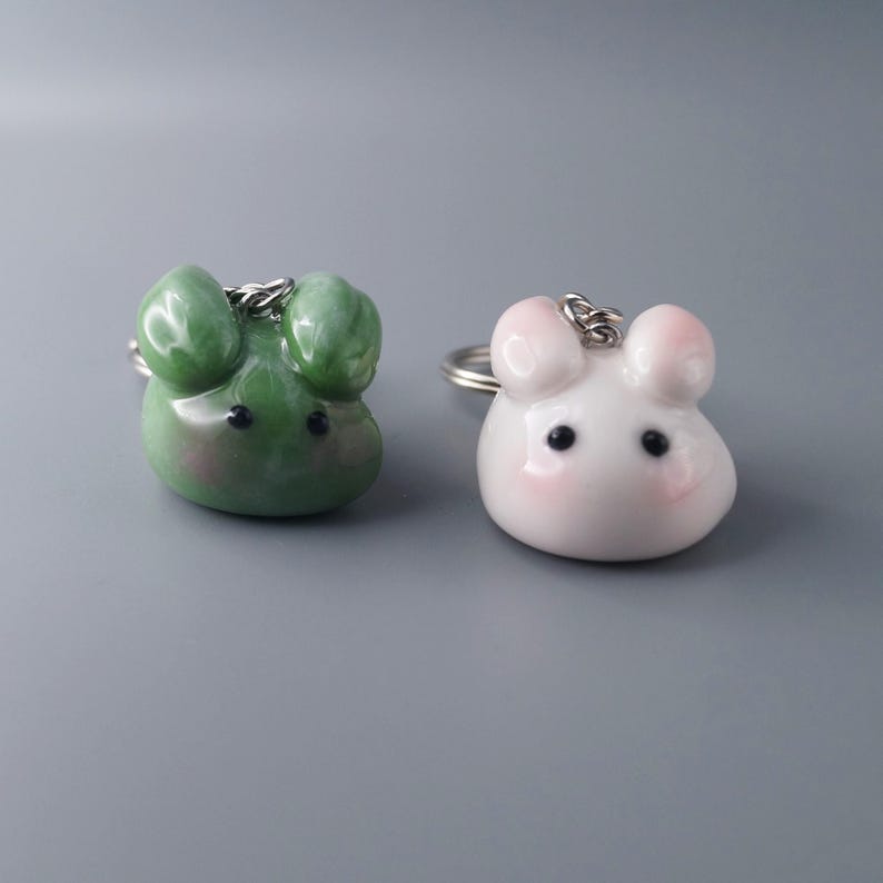 May include: Two keychains shaped like cartoon mice. One is green with black eyes, the other is white with pink accents. Both have silver key rings attached.
