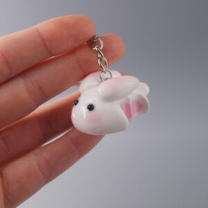May include: A white bunny keychain with pink accents on the ears and inner paws. The bunny has black eyes and rosy cheeks. The keychain is attached to a silver chain.