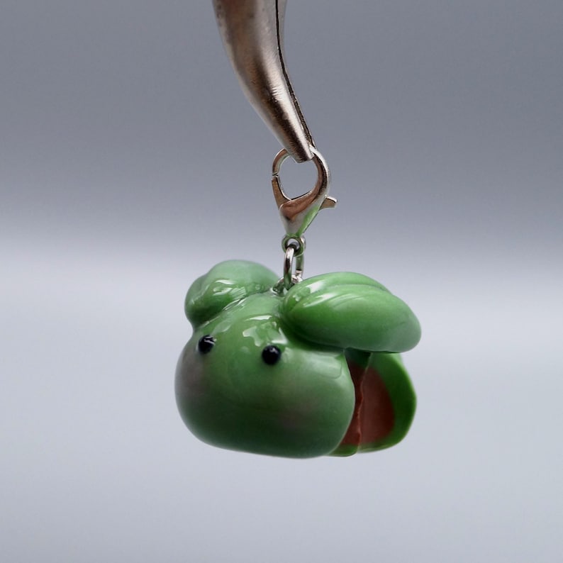 May include: A green charm shaped like an avocado, with a silver clasp. The avocado has a glossy finish, with a brown pit visible. The charm is held by a silver tool against a light gray background. The charm is a miniature, handcrafted item.