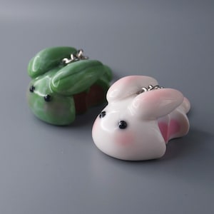 May include: Two bunny-shaped keychains. One is white with pink accents, the other is green. Both have black eyes and silver-colored key rings. The keychains are lying on a gray surface.