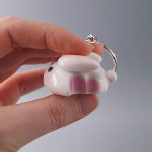 May include: A white and pink bunny keychain with a silver keyring. The bunny has pink ears, cheeks, and inner legs. The keychain is held in a person's hand against a gray background. The bunny is a cute, cartoon-like design.