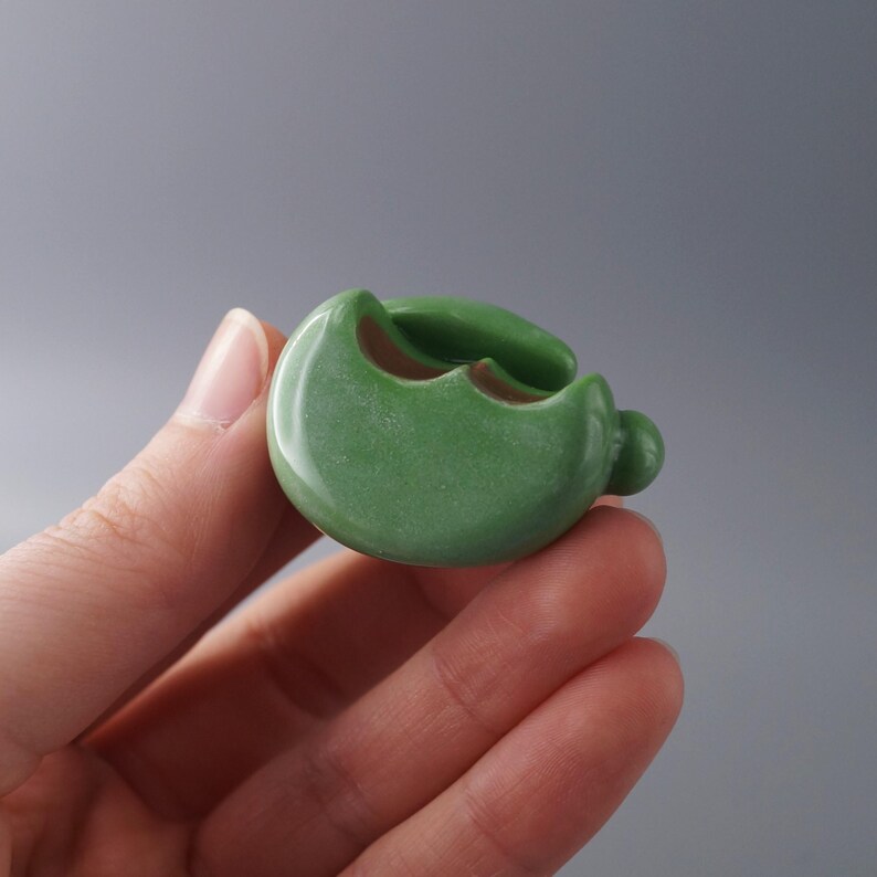 May include: A small, round, green ceramic container with a unique, organic shape. The container has a smooth, glossy finish and a small, rounded protrusion on one side. The interior is a contrasting brown color. The container is held in a hand.