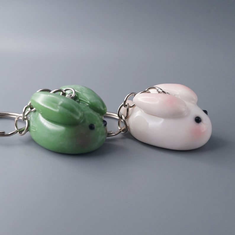 May include: Two bunny-shaped keychains. One is a glossy green, the other is white with pink accents on the ears and cheeks. Both have black eyes and silver key rings. The keychains are likely made of ceramic or a similar material.