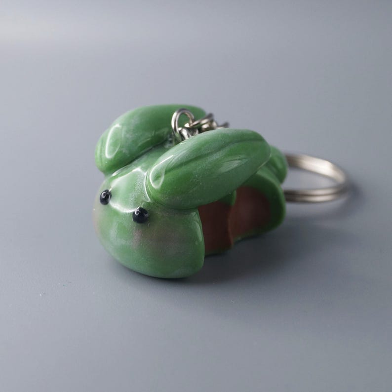 May include: A green frog-shaped keychain with a silver keyring. The frog has a glossy finish, black eyes, and a light brown underside. The keychain is made of a smooth, possibly polymer clay material.