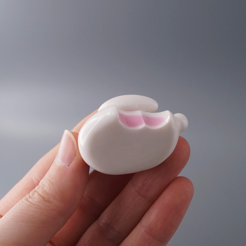 May include: A white ceramic bunny-shaped object with pink accents on the inside of the mouth. The bunny has a rounded body and a small, raised ear. The object is held in a hand against a gray background.