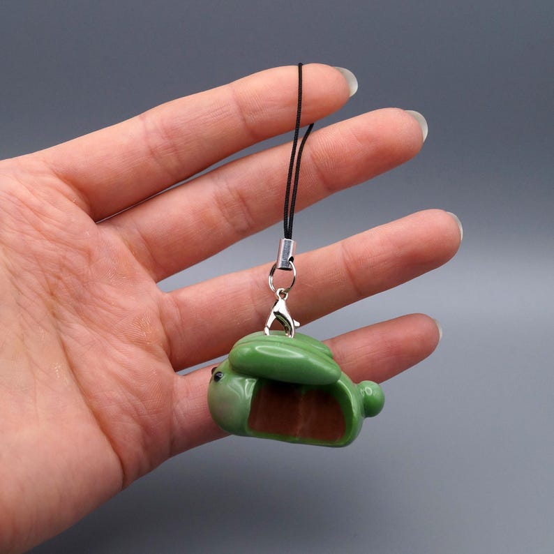 May include: A green and brown frog-shaped charm with a black cord and silver-colored clasp. The frog has a detailed design and a small, round body. The charm is held in a hand against a gray background.