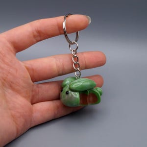 May include: A green and brown keychain featuring a small, stylized bee. The bee-shaped charm is attached to a silver metal keyring and chain. The keychain is held against a gray background.