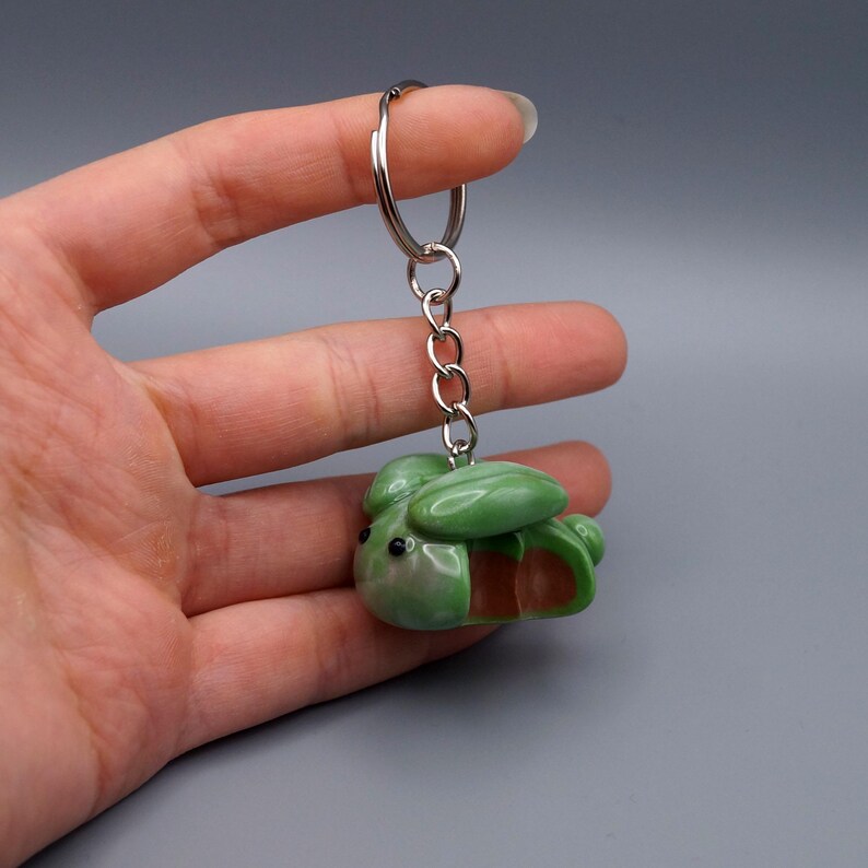 May include: A green and brown keychain featuring a small, stylized bee. The bee-shaped charm is attached to a silver metal keyring and chain. The keychain is held against a gray background.
