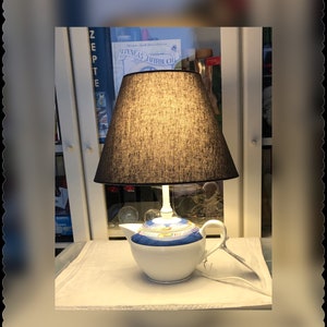 Könnte beinhalten: Eine weiße Teekannenlampe mit blauem und goldenem Design. Die Lampe hat einen schwarzen Stofflampenschirm und ein weißes Kabel.