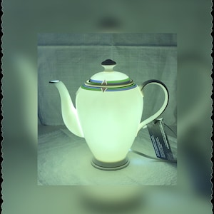 Könnte beinhalten: Eine weiße Teekanne aus Keramik mit grünen, blauen und braunen Streifen um den oberen Rand. Die Teekanne hat ein Licht im Inneren, das das Innere beleuchtet.
