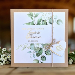 Könnte beinhalten: Eine quadratische Hochzeitseinladung mit einem Kraftpapierrand und einer weißen Mitte. Die Einladung zeigt goldene Schriftzüge mit den Namen "Leonie & Francesco" und dem Datum "23.04.2026". Sie ist mit grünem Laub und einem herzförmigen Anhänger verziert.