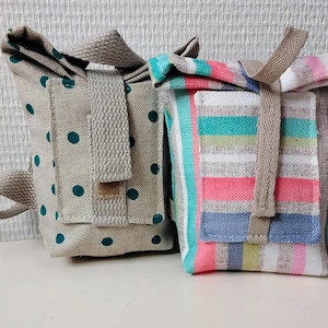Op de afbeelding: Twee kleine stoffen tassen. Eén is beige met teal stippen, de andere is gestreept met roze, blauw, groen en wit. Beide hebben een klepsluiting en een beige band. De tassen staan op een wit oppervlak.
