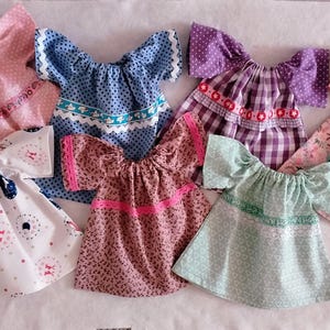 Könnte beinhalten: Eine Sammlung handgefertigter Puppenkleider in verschiedenen Farben und Mustern. Die Kleider weisen Details wie Blumenmuster, Tupfen und Spitzentrimm auf. Die Farbpalette umfasst Rosa, Blau, Lila und Weiß und zeigt eine Vielzahl von Designs.