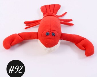 May include: Red plush lobster toy with white fur on the underside and blue button eyes. The lobster is lying on its back with its claws outstretched.