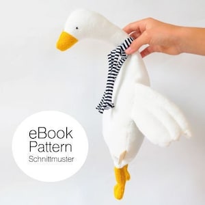 Könnte beinhalten: Weißes Plüsch-Gänse-Spielzeug mit gelbem Schnabel und Füßen, das einen schwarz-weiß gestreiften Schal trägt. Das Spielzeug wird von einer Hand gehalten. Der Text "eBook Pattern Schnittmuster" ist im Bild sichtbar.