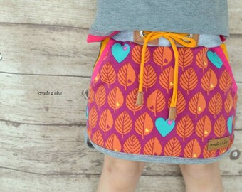 May include: A pink skirt with a colorful leaf pattern and a yellow drawstring. The skirt has two pockets on the sides and a small heart-shaped patch with the text "amelie & luise" on the right side.