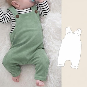 May include: A light green corduroy baby overall with wooden buttons on the straps. The baby overall is shown on a white fluffy surface. A black and white striped long-sleeve shirt is visible underneath the overall.