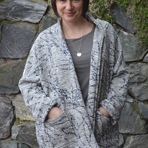 May include: A grey open-front cardigan with a navy blue floral pattern. The cardigan has two large pockets and is worn over a grey top and blue jeans. The background is a stone wall.