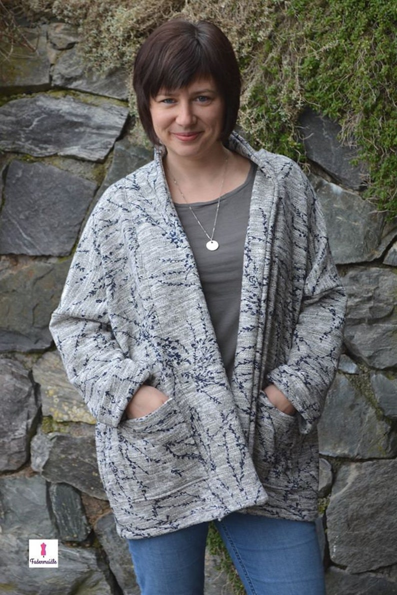 May include: A grey open-front cardigan with a navy blue floral pattern. The cardigan has two large pockets and is worn over a grey top and blue jeans. The background is a stone wall.