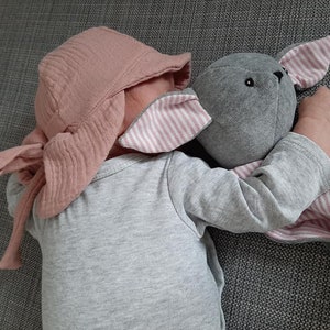 May include: A baby wearing a light pink bonnet and a gray onesie, cuddling a gray stuffed bunny with pink and white striped ears and a matching blanket. The bunny has black eyes.