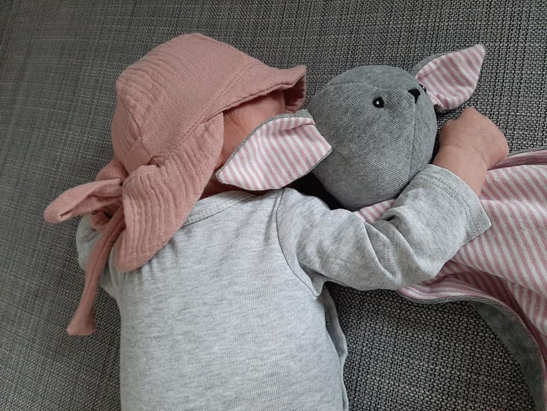 May include: A baby wearing a light pink bonnet and a gray onesie, cuddling a gray stuffed bunny with pink and white striped ears and a matching blanket. The bunny has black eyes.