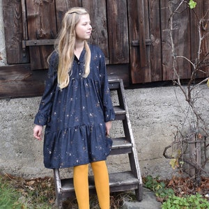 Puede incluir: Una persona joven lleva un vestido azul oscuro con estampado floral blanco y medias amarillas. Est&aacute; de pie en una escalera de madera frente a una puerta de granero de madera.