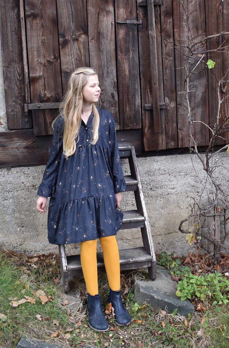 Puede incluir: Una persona joven lleva un vestido azul oscuro con estampado floral blanco y medias amarillas. Est&aacute; de pie en una escalera de madera frente a una puerta de granero de madera.