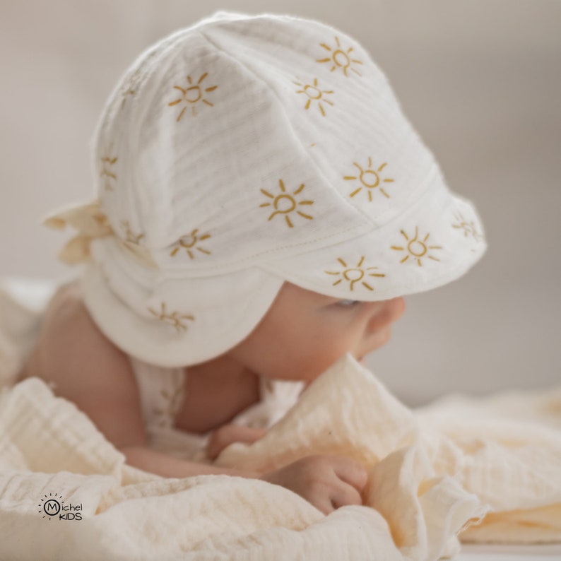 May include: A white baby sun hat with a brim and ear flaps, adorned with a repeating pattern of yellow suns. The hat is made of a soft, textured fabric. The image also shows a baby wearing the hat.