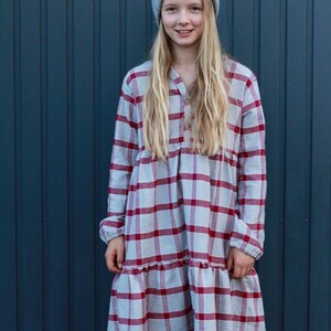 Puede incluir: Una persona joven con una boina gris y un vestido de cuadros rojos y blancos. El vestido tiene un dobladillo con volantes y mangas largas. La persona est&aacute; de pie frente a una pared azul.