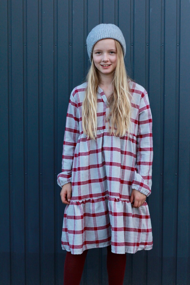 Puede incluir: Una persona joven con una boina gris y un vestido de cuadros rojos y blancos. El vestido tiene un dobladillo con volantes y mangas largas. La persona est&aacute; de pie frente a una pared azul.