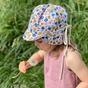 May include: A floral print sun hat with a brim, featuring blue, yellow, and pink flowers on a white background. The hat has cream-colored ties. The image also shows a pink dress and the text "MELA by Melanie Gieske".