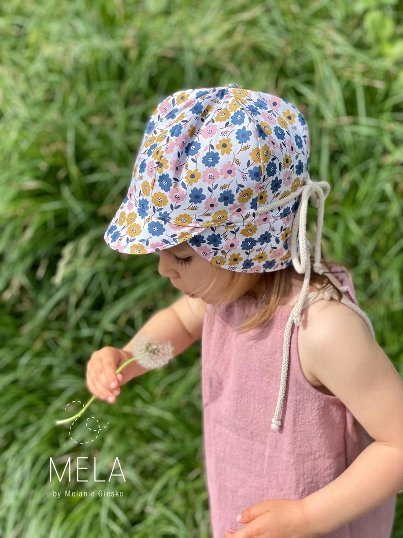 May include: A floral print sun hat with a brim, featuring blue, yellow, and pink flowers on a white background. The hat has cream-colored ties. The image also shows a pink dress and the text "MELA by Melanie Gieske".