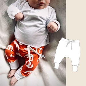 Puede incluir: Un bebé con una camisa blanca de manga larga y pantalones naranjas con un patrón blanco. Los pantalones tienen un cordón blanco. El bebé está sentado en una manta blanca. Un contorno blanco de un pantalón con un cordón se muestra en el lado derecho de la imagen.