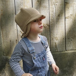 May include: A child wearing a tan-colored hat with ear flaps and a brim. The child is also wearing a striped long-sleeve shirt and blue pinstripe overalls. The background is a weathered wooden fence.