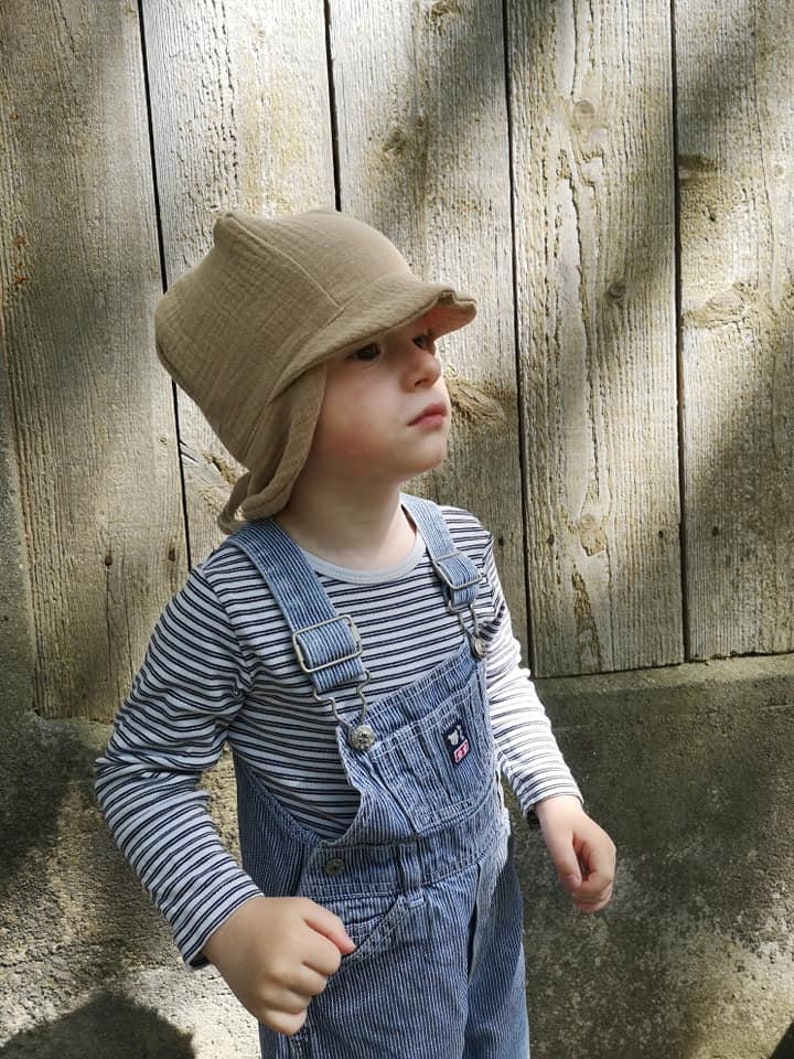 May include: A child wearing a tan-colored hat with ear flaps and a brim. The child is also wearing a striped long-sleeve shirt and blue pinstripe overalls. The background is a weathered wooden fence.
