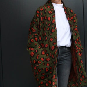 May include: An olive green coat featuring a red leopard print design. The coat has large pockets and is worn over a white top and dark grey jeans. A fashionable outerwear garment.