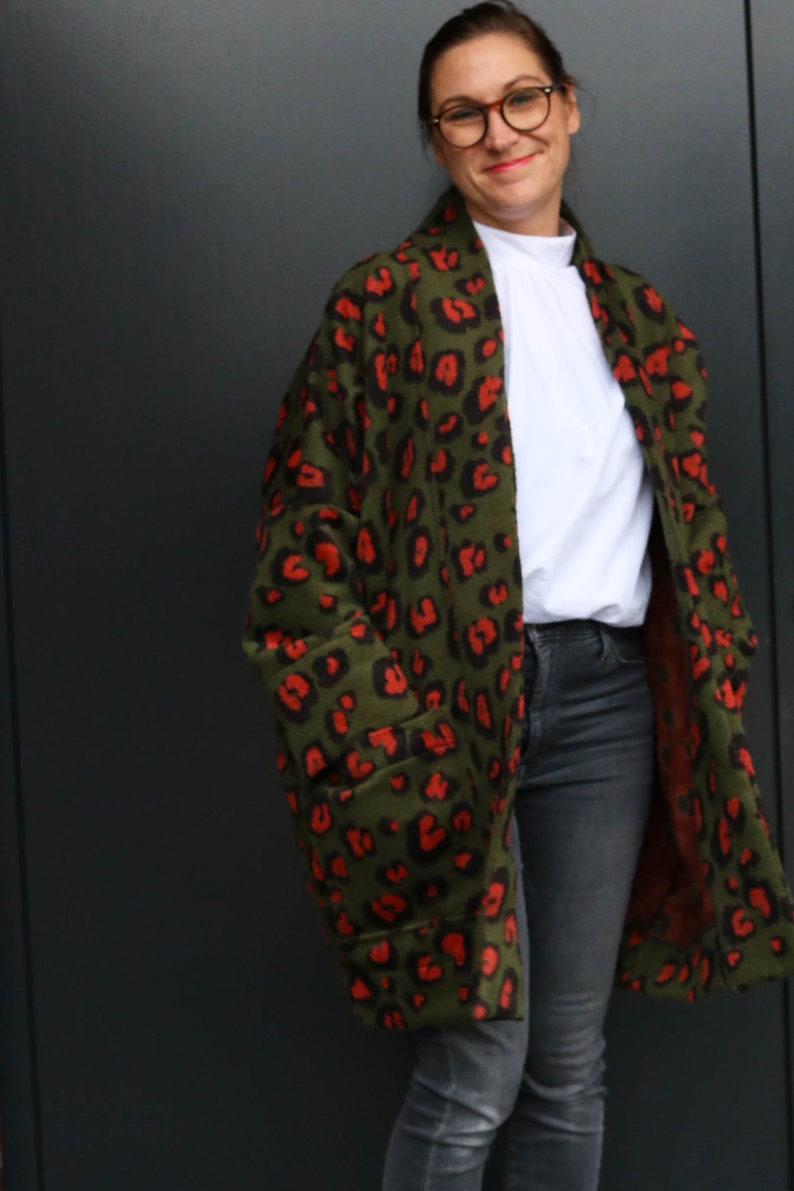 May include: An olive green coat featuring a red leopard print design. The coat has large pockets and is worn over a white top and dark grey jeans. A fashionable outerwear garment.