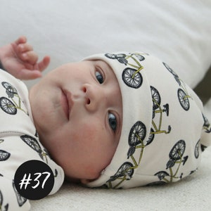 Könnte beinhalten: Eine weiße Baby-Mütze mit einem schwarzen und gelben Fahrrad-Druck. Die Mütze hat einen umgeschlagenen Rand und ist aus weichem, atmungsaktivem Stoff. #37